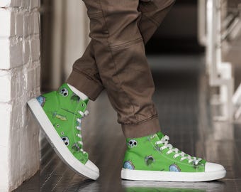 Op de afbeelding: Hoge canvas sneakers met een felgroene achtergrond en een herhaald patroon van cartoon schedels, klokken en andere graphics. De schoenen hebben witte veters, een witte zool en een witte neus. De schoenen worden gedragen door een persoon die een bruine broek draagt.