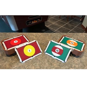 May include: Four miniature pool table ball sets, each with a different number. The balls are red, yellow, and green with white borders. Each set is in a brown container. The numbers are 1, 3, 13, and 15.