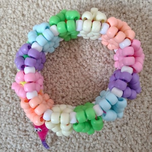 May include: A colorful beaded bracelet in a circular shape. The bracelet features a variety of pastel colors, including pink, orange, yellow, green, blue, and purple. A small, pink worm with googly eyes is attached to the bracelet.