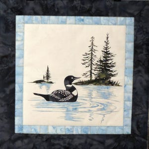 May include: A square quilt featuring a loon swimming in a lake with pine trees in the background. The loon is black and white, and the water is light blue. The quilt has a light blue border and a dark blue outer border.