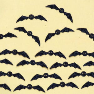 May include: A collection of dark blue bat-shaped embellishments with a small, round, black center. The bats are arranged in a triangular pattern against a light yellow background, suitable for Halloween crafts.