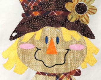 May include: A cheerful scarecrow face applique with a brown hat, yellow hair, and a smiling face. The hat features a plaid patch and a decorative flower. The scarecrow has blue eyes, a pink nose, and a plaid scarf.