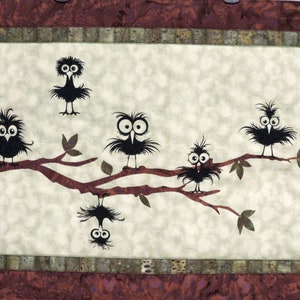 May include: A whimsical quilt featuring five black birds with large eyes perched on a brown branch against a cream background. The birds have a textured, fluffy appearance and are arranged in a playful, asymmetrical pattern.