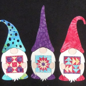 May include: Three gnomes with different colored hats and patterned fabric holding patchwork squares. The gnome on the left has a blue and white hat, the middle gnome has a purple hat, and the gnome on the right has a red hat.