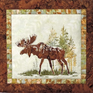 May include: A square fabric wall hanging featuring a central panel with a brown moose silhouette standing in a forest of green and tan trees. The panel is framed by a border of small, multicolored squares and a wide, dark brown outer fabric border.
