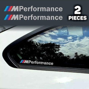 Pode incluir: Um conjunto de dois autocolantes MPerformance com as icónicas riscas inclinadas em azul, roxo e vermelho, seguidas do texto MPerformance em branco. Os autocolantes estão aplicados no vidro lateral traseiro de um veículo branco.
