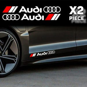 Peut inclure: Voiture noire avec des autocollants Audi blancs et rouges. Les autocollants incluent le logo et le texte Audi, avec une bande rouge. L'image montre la roue et le panneau latéral de la voiture, avec le texte "X2 PIECE" en blanc.