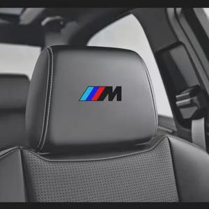 May include: Black car seat headrest with a white stitched border. The headrest features the BMW M logo in blue, red, and black. The seat has a perforated leather design.