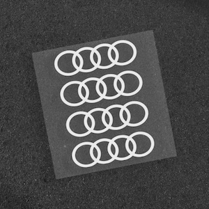 May include: A sheet of white vinyl decals featuring the Audi logo, composed of four interlocking rings. The decals are arranged in a grid on a transparent backing, set against a dark, textured background.