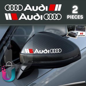 以下が含まれることがあります： 白のアウディロゴステッカーに赤いアクセントと「Audi」の文字が表示されています。画像は車のミラーにステッカーが貼られている様子です。「2 PIECES」の文字も表示されています。ミラーはカーボンファイバーパターンです。