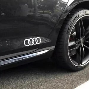 May include: A black car with a white Audi emblem on the side. The car has a black tyre with a Michelin logo and a black rim. The car is parked on a grey surface.