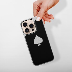 May include: A black smartphone case with a silver spade symbol. The case has a clear border and a gold camera lens protector. The phone is held by a hand with white nail polish against a white background.