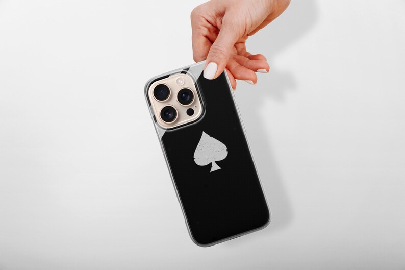 May include: A black smartphone case with a silver spade symbol. The case has a clear border and a gold camera lens protector. The phone is held by a hand with white nail polish against a white background.