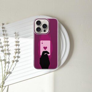 May include: A magenta phone case featuring a playing card design. The case displays a silhouette of a hand holding the Ace of Hearts card. The phone case is on a white plate with a sprig of dried flowers.