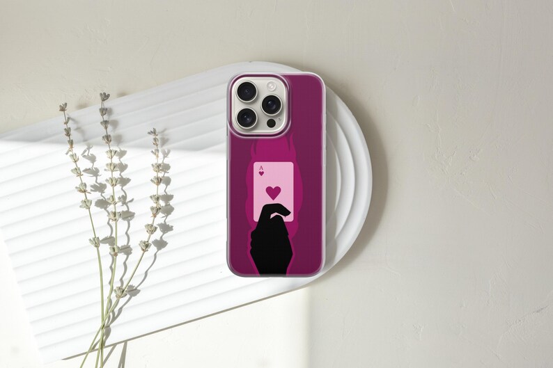 May include: A magenta phone case featuring a playing card design. The case displays a silhouette of a hand holding the Ace of Hearts card. The phone case is on a white plate with a sprig of dried flowers.