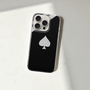 May include: A black smartphone case with a white spade symbol. The case has a clear border and a glossy finish. The phone's camera lenses are visible at the top. The case is lying on a white surface.