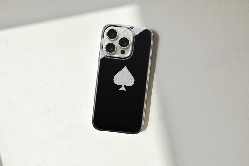 May include: A black smartphone case with a white spade symbol. The case has a clear border and a glossy finish. The phone's camera lenses are visible at the top. The case is lying on a white surface.