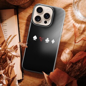 May include: A black phone case featuring playing card suit symbols: spade, heart, club, and diamond. The case is on a wooden surface with a book, dried leaves, and a glass of water. The phone case has a clear border.