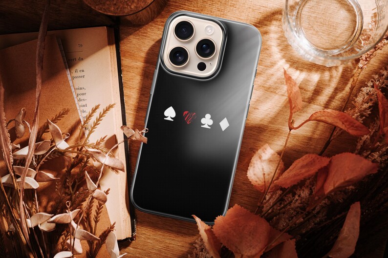 May include: A black phone case featuring playing card suit symbols: spade, heart, club, and diamond. The case is on a wooden surface with a book, dried leaves, and a glass of water. The phone case has a clear border.