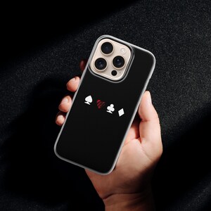 May include: Black phone case with a playing card suit design. The case features white spade, red heart, white club, and white diamond symbols. The phone case is held in a hand against a dark background.