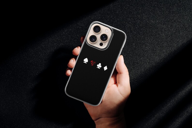 May include: Black phone case with a playing card suit design. The case features white spade, red heart, white club, and white diamond symbols. The phone case is held in a hand against a dark background.