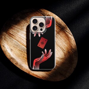 May include: A clear phone case featuring a black background with a red playing card and red hands. The phone case is resting on a wooden, oval-shaped tray, with a dark background.
