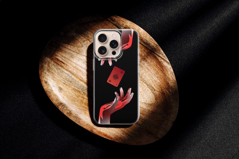 May include: A clear phone case featuring a black background with a red playing card and red hands. The phone case is resting on a wooden, oval-shaped tray, with a dark background.