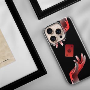 May include: Black phone case featuring a red and white illustration of hands and a playing card. The card is the Ace of Spades. The phone case is next to a framed piece of art.