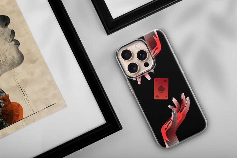 May include: Black phone case featuring a red and white illustration of hands and a playing card. The card is the Ace of Spades. The phone case is next to a framed piece of art.