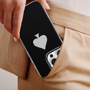 May include: A black phone case with a white spade symbol is partially visible in a pocket. The case has a clear border and is set against a tan background. The phone's camera lenses are also visible.
