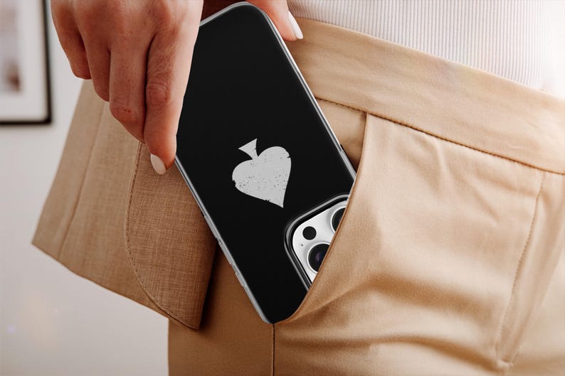 May include: A black phone case with a white spade symbol is partially visible in a pocket. The case has a clear border and is set against a tan background. The phone's camera lenses are also visible.
