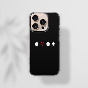 May include: Black smartphone case with a clear border. The case features white spade, club, and diamond symbols, and a red heart symbol. The phone case is on a white surface with a shadow.
