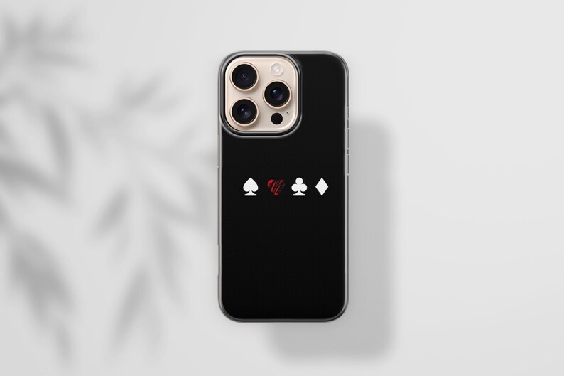 May include: Black smartphone case with a clear border. The case features white spade, club, and diamond symbols, and a red heart symbol. The phone case is on a white surface with a shadow.
