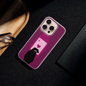 May include: A magenta phone case featuring a playing card design with a heart symbol. The case is on a black surface, next to a wooden tray, glasses, and a black notebook.