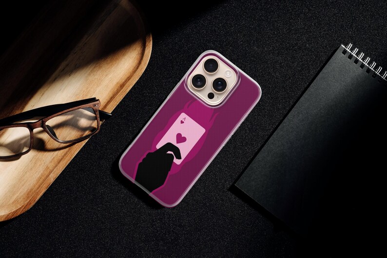 May include: A magenta phone case featuring a playing card design with a heart symbol. The case is on a black surface, next to a wooden tray, glasses, and a black notebook.