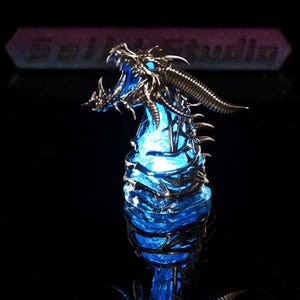 Könnte beinhalten: Eine metallische Drachenkopfskulptur mit offenem Maul, aus deren Innerem ein leuchtend blaues Licht strahlt. Die Basis ist mit komplizierten Metalldrahtdetails umwickelt. Der Text Saili Studio ist im Hintergrund zu sehen.