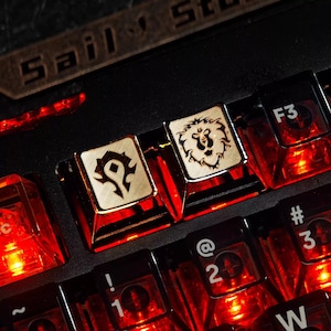 Può includere: Primo piano di una tastiera retroilluminata con tasti personalizzati. Due tasti presentano i simboli dell'Orda e dell'Alleanza di World of Warcraft. La tastiera ha un'illuminazione rossa e il testo "Sail" è visibile.