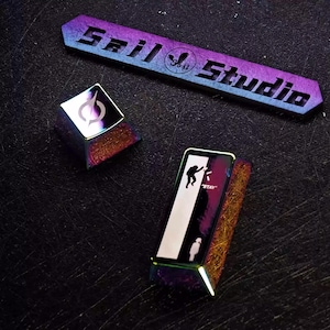 Può includere: Tasti per tastiera iridescenti e una lunga barra con la scritta "Sril Studio". I tasti hanno un design geometrico e la silhouette di una persona. La barra è un gradiente di blu, viola e oro.