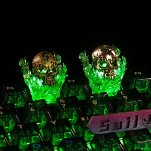 Könnte beinhalten: Zwei goldfarbene Schädel-Tastenkappen mit grünen LED-Flammen. Die Tastenkappen befinden sich auf einer Tastatur mit grüner Hintergrundbeleuchtung. Dunkler Hintergrund.