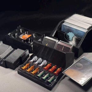 Pode incluir: Um organizador de jogos de tabuleiro em plástico preto com vários compartimentos para baralhos e fichas. O conjunto inclui ranhuras para cartas com a etiqueta Intrige, pilhas de cartas brancas e pequenas figuras metálicas em dourado, prateado e verde.