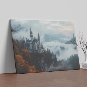 May include: A canvas print depicting a fairytale castle in a mountainous, misty landscape. The castle has multiple towers and spires. Autumnal colours of orange and red surround the castle. A white vase with branches is on a wooden surface.