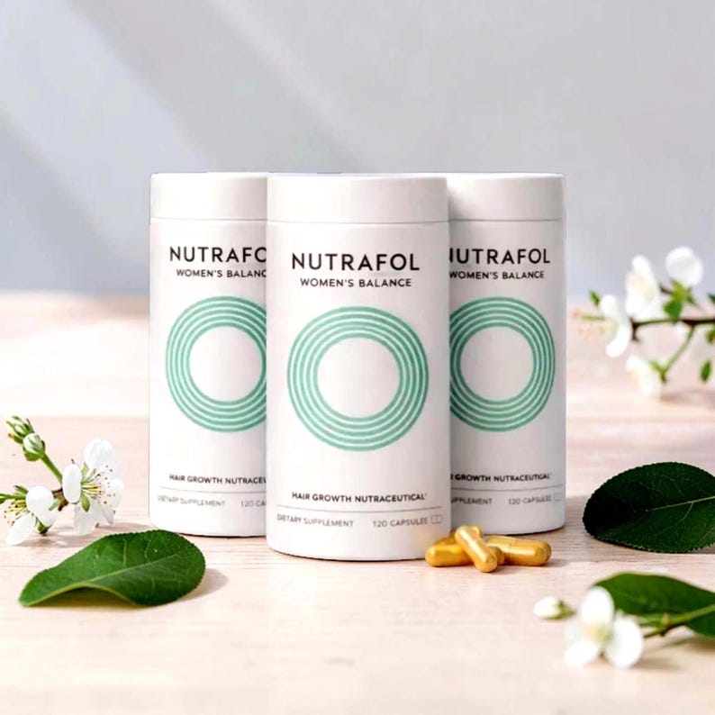 Puede incluir: Tres envases cil&iacute;ndricos blancos de suplemento para el crecimiento del cabello Nutrafol Women's Balance. Cada envase tiene un dise&ntilde;o circular turquesa y el texto "Nutrafol Women's Balance". C&aacute;psulas doradas frente a los envases. Flores blancas y hojas verdes en el fondo.