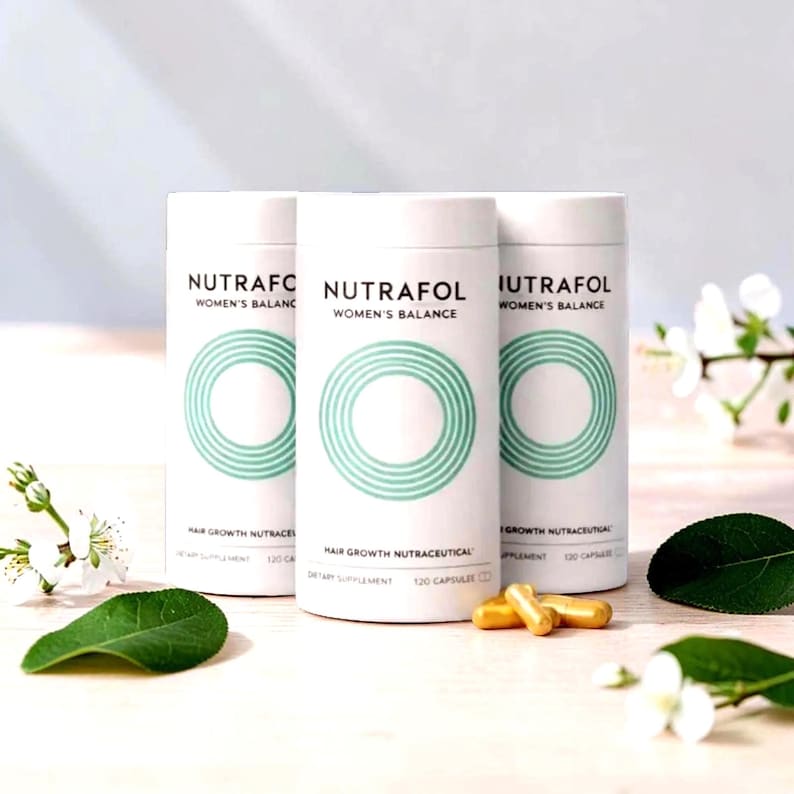 Puede incluir: Tres envases cil&iacute;ndricos blancos de "Nutrafol Women's Balance", nutrac&eacute;uticos para el crecimiento del cabello. Cada envase tiene un dise&ntilde;o circular turquesa y el texto "Hair Growth Nutraceutical" y "120 Capsules". Varias c&aacute;psulas doradas est&aacute;n delante de los envases.