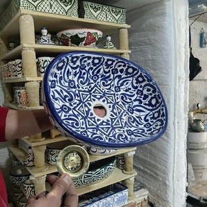 May include: An oval ceramic sink with a blue and white floral pattern is held in front of a kiln. A brass drain is held in front of the sink. Other ceramic pieces are on shelves in the background.