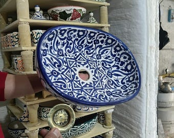 May include: An oval ceramic sink with a blue and white floral pattern is held in front of a kiln. A brass drain is held in front of the sink. Other ceramic pieces are on shelves in the background.