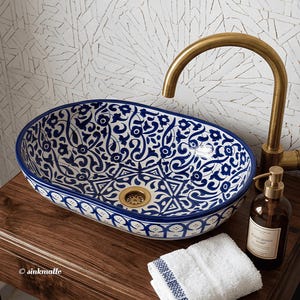 May include: An oval ceramic vessel sink featuring a blue and white floral pattern, paired with a curved brass faucet and a matching brass drain. A brown glass soap dispenser and a folded white towel with blue trim are placed nearby.