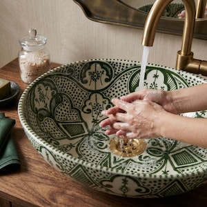 Peut inclure: Une vasque ronde en céramique à fond blanc ornée de motifs complexes peints à la main en vert foncé. Le lavabo est équipé d'une bonde dorée et d'un robinet courbé en laiton. Le design présente des motifs floraux et géométriques symétriques.