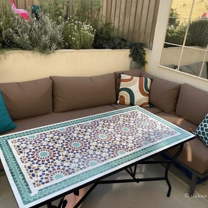 May include: An outdoor sectional sofa with brown cushions and teal throw pillows. A rectangular coffee table with a mosaic tile top in shades of blue, brown, and gold sits in front of the sofa. The table is supported by a black metal frame.