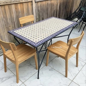 May include: An outdoor dining set comprising a rectangular table with a patterned tile top and a black metal frame. Three wooden chairs surround the table. The table's surface has a white background with a blue dot pattern.