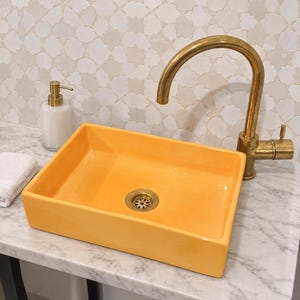 May include: A rectangular bright yellow ceramic vessel sink with a gold-toned drain, paired with a curved brass faucet. A white soap dispenser with a gold pump and a folded white cloth are positioned nearby on a white marble countertop.